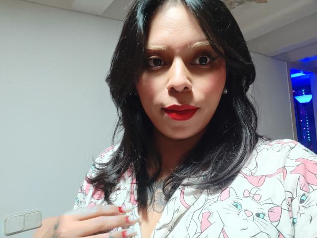 Close-up of Chili, a woman with long black hair, red lipstick, and a white floral shirt. She gazes directly at the camera, hand on chest, intense sexual tension. Bright lighting.