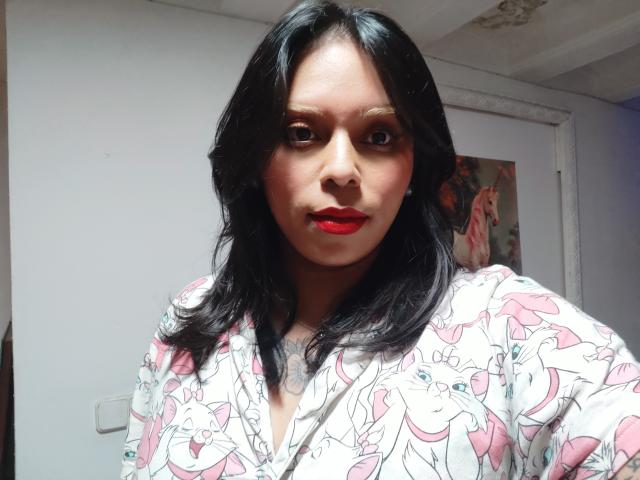 Close-up of Chili, a woman with dark, wavy hair and red lipstick, wearing a white shirt with pink floral patterns. Strong lighting creates shadows on her face, highlighting her intense gaze.