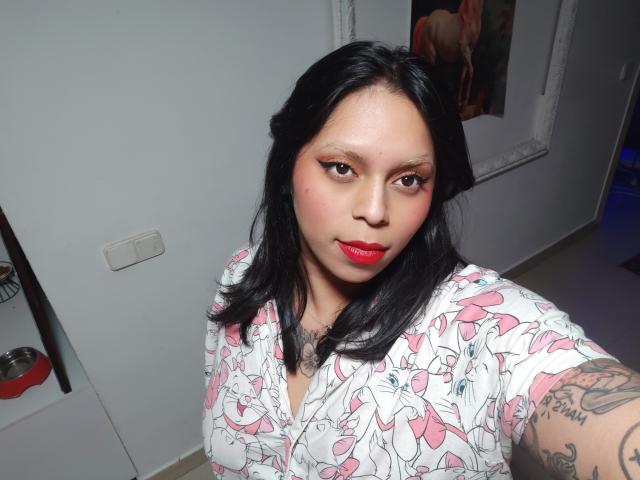 Photograph of a plus-sized woman with medium brown skin, black hair, red lipstick, and a white dress with pink and red abstract patterns. She's looking directly at the camera with a slight smirk. Her left arm has a visible tattoo. Bright lighting highlights her face and red lips. Background includes a white wall and a door.