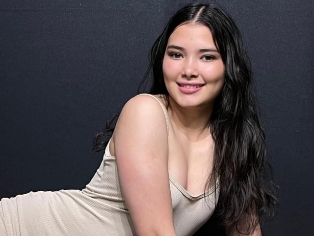 Photographie de LizzieSweets, une femme asiatique aux longs cheveux noirs, portant un débardeur blanc moulant, penchée en avant. Un sourire joueur, mettant en valeur sa poitrine et son allure séduisante. Fond noir.