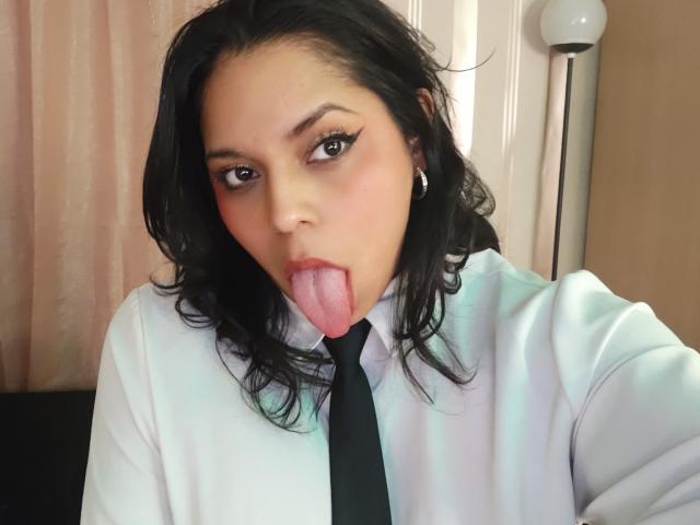 Photograph of a young woman, Chili, with black hair, sticking her tongue out while wearing a white shirt and black tie. Her eyes are half-open, creating a suggestive expression.