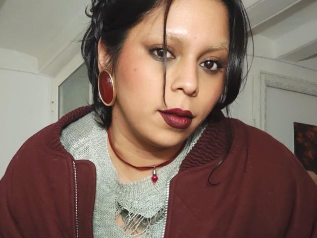 Close-up of Chili with medium skin tone, black hair, and dark red lipstick. Wearing a maroon hoodie over a lace shirt, large red hoop earrings, and a red pendant necklace. Fucking intense gaze.
