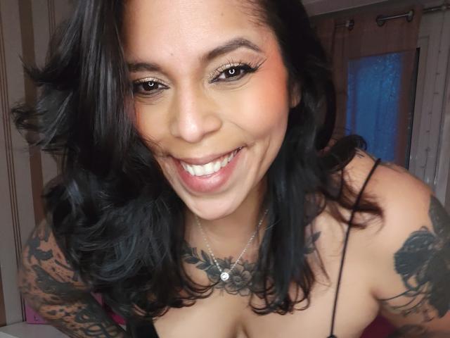 Close-up of Chili with black wavy hair, smiling widely, showing off white teeth. She has dark tattoos on her arms, wears a thin-strapped top, and a necklace. Fucking hot.