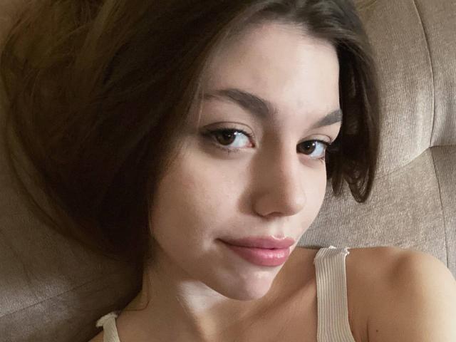 Close-up photo of StacyStripling with fair skin and dark brown hair, lying on a beige couch. She has a slight smile, dark eyes, and is wearing a white strap top. Sexual tension.