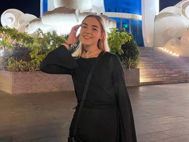 RoxyFoxy-hot, smiling with hand on cheek, wearing black long-sleeve dress and necklace, standing in front of illuminated building with green bushes, bright lights, and modern architecture. Fucking cute.