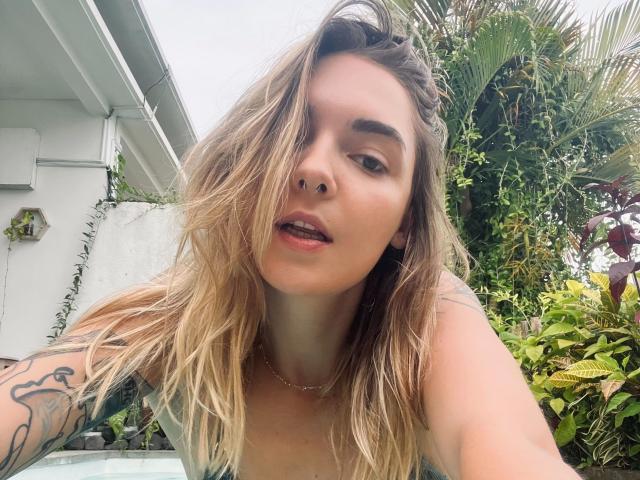 Gros plan de RoxyFoxy : cheveux blonds et ondulés, piercing au nez, bras tatoué, en extérieur, plantes vertes luxuriantes, mur blanc en arrière-plan. fond, lumière du soleil, bouche légèrement ouverte, décontracté, sexy, regard direct, ambiance estivale..