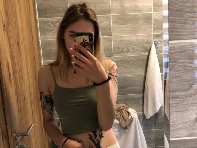 Selfie de RoxyFoxy, muito atraente, em um banheiro, segurando um celular com o braço tatuado, vestindo uma cropped verde, revelando a barriga plana. Tatuagens pretas na região da virilha, parede com azulejos cinzas, toalha branca pendurada. Muito quente.