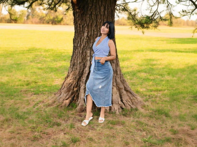 LucyDaniels est appuyée contre un tronc d'arbre épais dans un champ herbeux baigné de soleil. Elle porte un haut bleu à motifs et une jupe en jean, ainsi que des chaussures plates blanches. Ses cheveux noirs tombent librement. Une ambiance vraiment détendue..