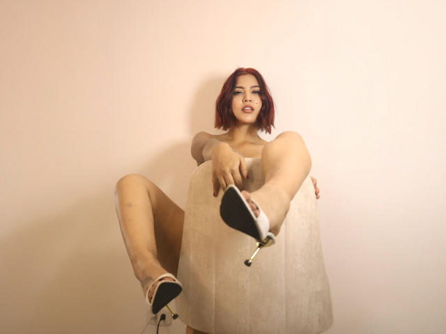 LucyDaniels, met rood haar, leunt tegen een witte muur, met gespreide benen, en draagt een beige jurk en zwarte hakken. Haar hand rust op haar dij, wat een seksuele spanning uitstraalt. Heldere verlichting accentueert haar.