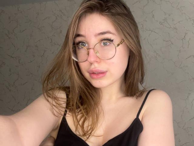 RobbieElsa, met een lichte huid en blauwe ogen, draagt ronde brillen en een zwart topje met spaghettibandjes, met lichtroze lippen. lippen, bruin haar en een gekanteld hoofd, die seksuele spanning uitstraalden. Achtergrond met een gebarsten muur..