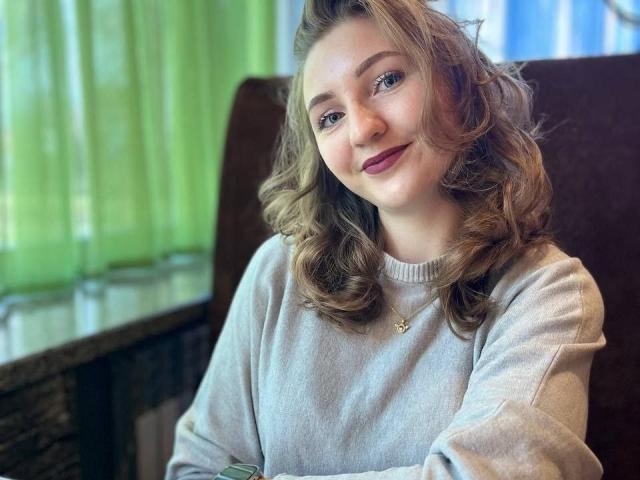 SamuelaBella has wavy brown hair, wearing a white sweater, sitting with a slight smile. Soft light from green curtains highlights her fair skin. Background is blurred, focusing on her. Fucking cute.