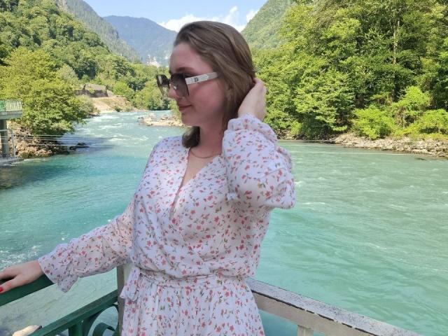 SamuelaBella, with medium-length brown hair, wearing a white floral dress, stands on a riverboat, hand in hair, looking right. Blue river, lush green trees, mountains in background. Fucking beautiful view.
