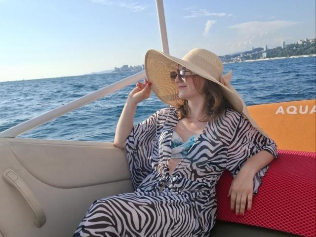 SamuelaBella sits on a boat, wearing a zebra-print dress and wide-brimmed straw hat, holding the brim with her right hand, blue ocean and city skyline in the background. Fucking hot.