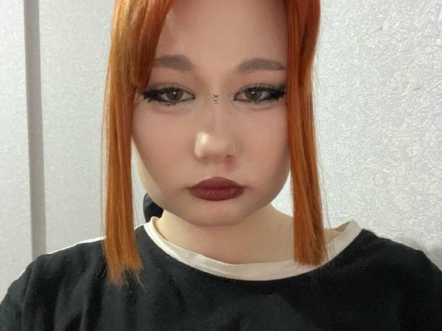 Fotografia ravvicinata di AngelaRamal con capelli rossi lisci, lunghi fino alle spalle, eyeliner nero intenso, e rossetto marrone scuro. Asfalto nero.
lei guarda direttamente la telecamera con un'espressione neutra.