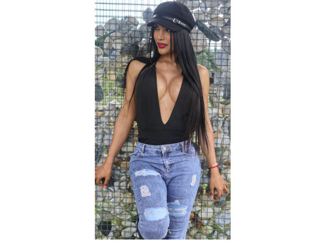 Vanezzattss com cabelos pretos longos, blusa preta com decote em V e calças jeans azuis rasgadas, em pé em frente a uma parede de tela. Ela usa um boné preto, batom vermelho e uma pulseira vermelha. Incrivelmente sexy..