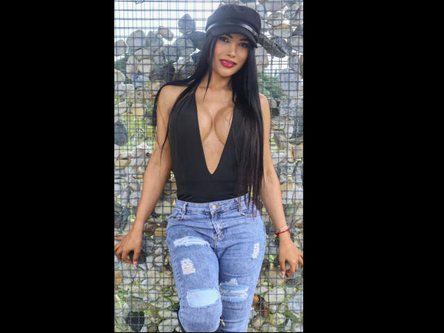 Vanezzattss con cabello negro largo, una blusa negra de cuello en V pronunciado y jeans azules rotos, usando una gorra negra, de pie contra pared con azulejos, mirando seductoramente a la cámara, con los labios entreabiertos..