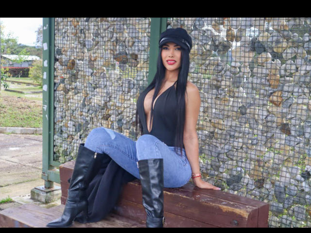 Una donna con lunghi capelli neri, rossetto rosso e cappello nero, seduta su una panca di legno, indossa una maglia nera che lascia intravedere scollatura, jeans blu e stivali neri alti fino al ginocchio. Sfondo con muro di pietra. Incredibilmente sexy..