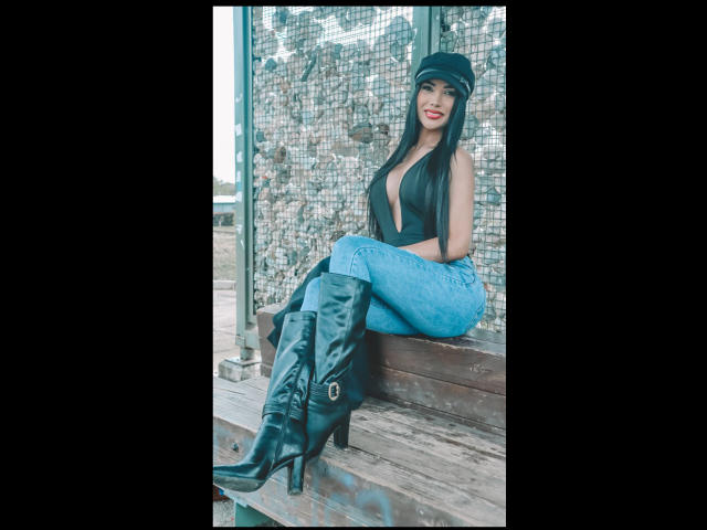 Vanezzattss, con el pelo largo y negro, está sentada en un banco de madera, vistiendo una camiseta negra, leggings de encaje azul y botas negras. Detalles de hebillas. Tiene una pose seductora, mostrando una sonrisa coqueta..