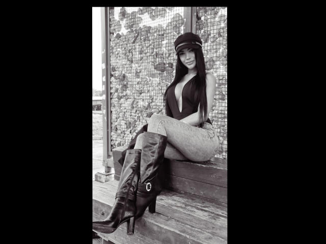 Foto em preto e branco de Vanezzattss sentada em um banco de madeira, usando um boné, uma blusa com decote em V profundo, calças justas e botas que chegam até a altura do joelho, com fivela. Pose sensual e confiante, olhando para a câmera..