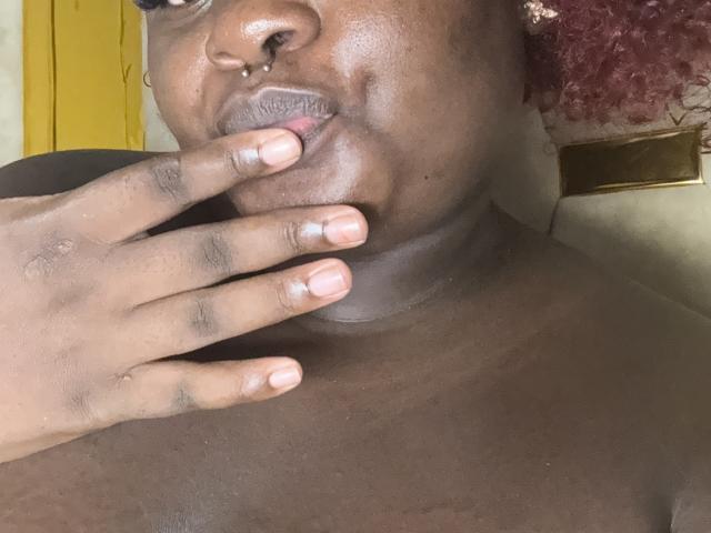 Primer plano del rostro de BigChocoTitiey, de piel oscura y brillante, con una mano cuyas puntas de los dedos cubren parcialmente la boca, ligeramente entreabierta. labios, fondo amarillo, tela roja estampada en el fondo, intensa tensión sexual, textura brillante..