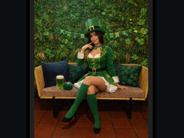 MindyLove sits on a wicker couch in a green leprechaun costume, legs crossed, hand near mouth, seductive smile. Green top hat, knee-high socks, cleavage exposed, beer on the couch.