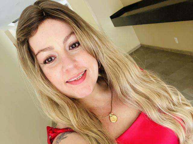 Nahaufnahme von SamanthaGold, die mit leuchtenden Augen lächelt, mit langen blonden Haaren, einem roten Oberteil und einer goldenen Halskette, wobei ein sichtbares Tattoo zu sehen ist. auf der linken Schulter, sexy, lässig, Innenaufnahme, weiches Licht, leichte Schatten..