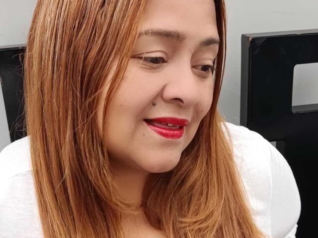 Primer plano de Monzerrat Boyer, una mujer de piel clara con cabello lacio de color castaño claro, lápiz labial rojo y una blusa blanca, que está mirando con una leve sonrisa insinuante, irradiando tensión sexual.