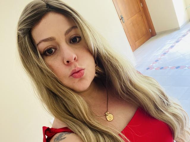 Autoritratto ravvicinato di SamanthaGold, una donna bionda con lunghi capelli, che indossa una maglietta rossa e una collana dorata, e guarda direttamente verso la fotocamera. la fotocamera, con le labbra leggermente dischiuse, un tatuaggio visibile sulla spalla destra..