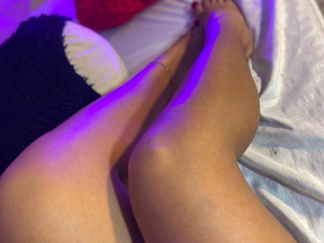Primer plano de las piernas de CelesteMorel, con una piel lisa y bronceada, cruzadas a la altura de los tobillos, mostrando un tobillero de plata. La luz de neón azul ilumina las piernas. El fondo incluye tela blanca y negra..
