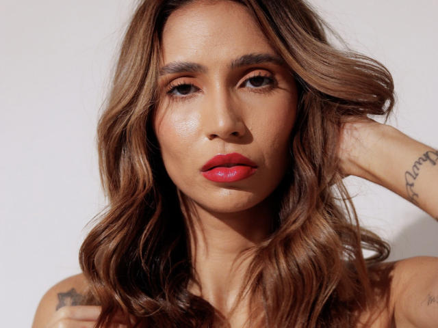 Photo en gros plan de BrasiNatural, avec des cheveux bruns ondulés, du rouge à lèvres rouge et un tatouage sur son bras droit. Elle regarde directement l'appareil photo, la main levée derrière sa tête. Extrêmement séduisant..