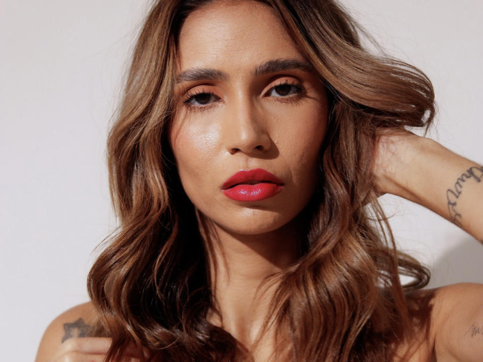 Photo en gros plan de BrasiNatural, avec des cheveux bruns ondulés, du rouge à lèvres rouge et un tatouage sur son bras droit. Elle regarde directement l'appareil photo, la main levée derrière sa tête. Extrêmement séduisant..