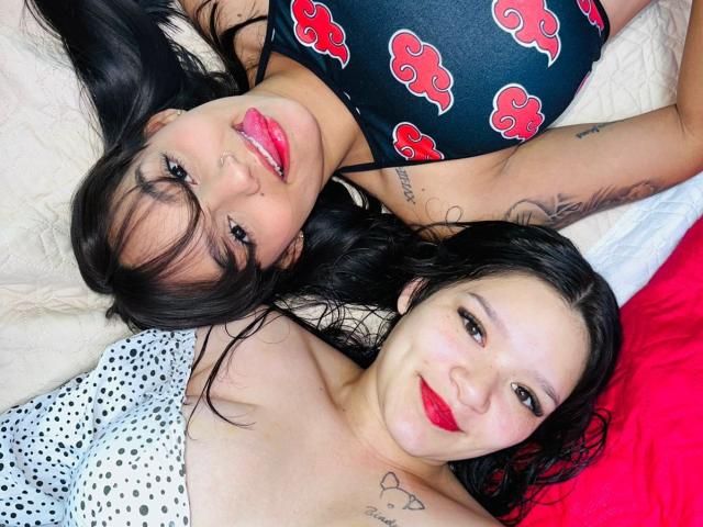 Zwei lesbische Frauen liegen auf einem Bett, die Gesichter nah beieinander, schwarzes Haar, roter Lippenstift, schwarzes Blumenkleid, weißes Top mit Punkten. Tattoos, sexuelle Spannung, inniger Blick, leuchtende Farben, rotes Kissen.