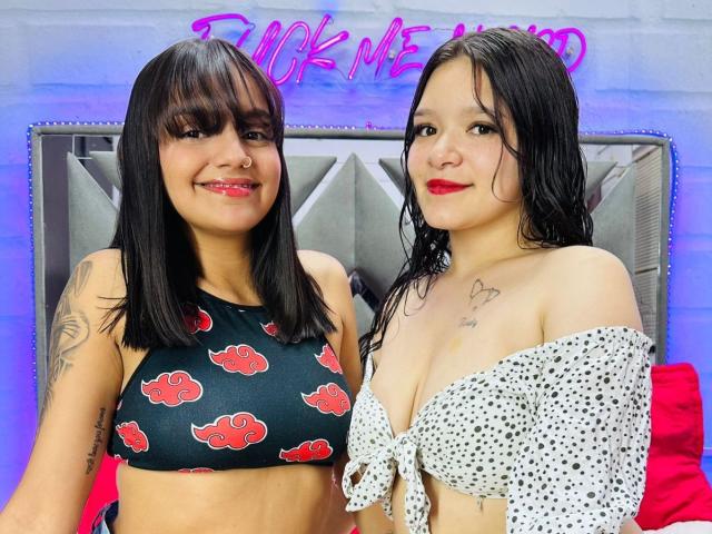 Two LesbiGeerShots, one with black crop top and red flowers, the other with white polka dot off-shoulder top, standing close, wet hair, tattoos, neon "FUCK ME HARD" sign behind.
