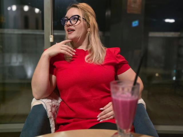 Foto van Angelina-Joline, aantrekkelijk met blond haar, die een rode blouse, zwarte broek en een bril draagt, en in een stoel zit. denken met hand op kin, roze drankje op de voorgrond, nachtelijke stad op de achtergrond.