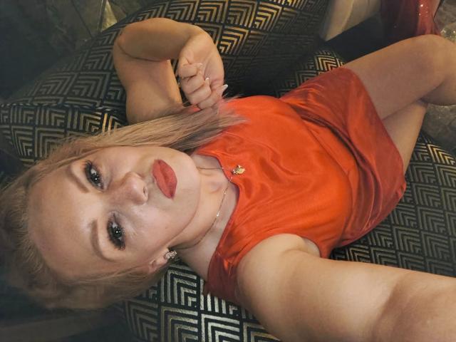 Angelina-Joline1-sexy ligt op een gestreept zwart en gouden bank, gekleed in een strakke rode jurk, en bijt verleidelijk op haar lip. Ze heeft lichtbruin haar, rode lippenstift en een gouden ketting. Heel aantrekkelijk..