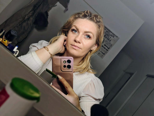 Fotografia de Angelina-Joline, atraente, loira, de olhos azuis, vestindo uma blusa branca, segurando um telefone rosa, olhando para o espelho, com a mão. no pescoço, sexy, levemente corada, quarto com pouca luz, xícara verde em primeiro plano..