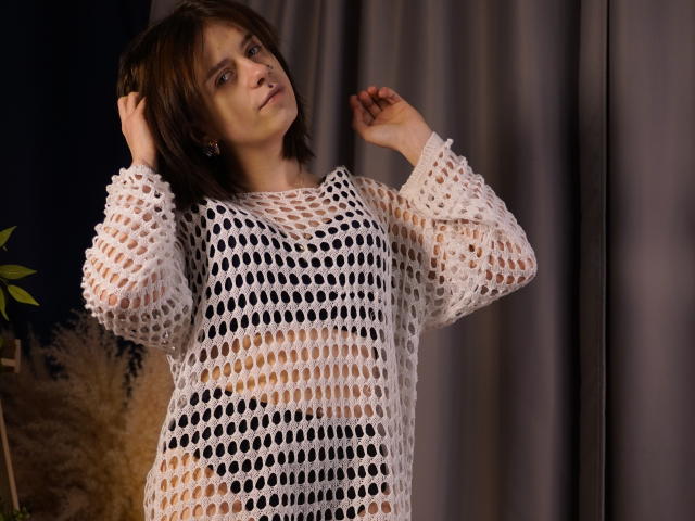 DejaSorley with shoulder-length dark hair, wearing a white, see-through knit sweater, stands against a dark curtain. Hands raised, expression sultry, showcasing a sexy, confident pose.