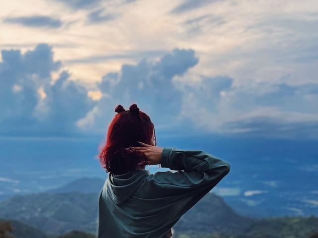 Photographie de SophieMaiden avec des cheveux roux, vue de dos, debout, une main sur la tête, regardant un ciel lumineux et nuageux. Ciel au-dessus des montagnes. Portant une veste sombre, les teintes bleues et grises prédominent..