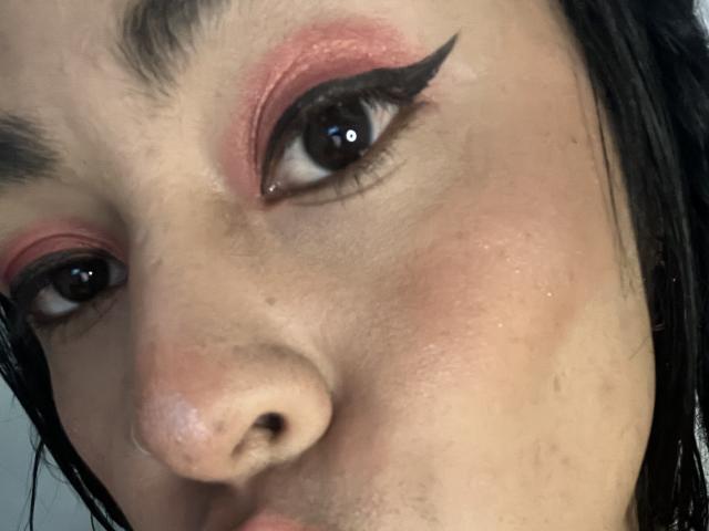 Close-up of StellaBeckker's face with intense eye makeup, thick black eyeliner, and pink eyeshadow. Her dark hair frames her face. Her expression is slightly parted lips, gazing directly.