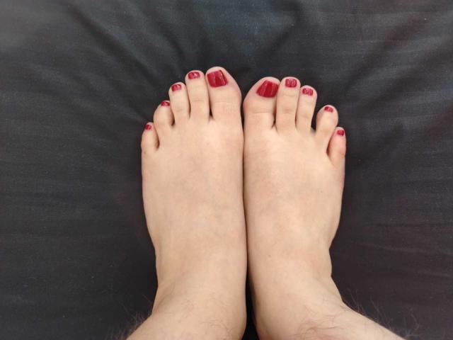 Close-up dos pés pálidos de TaylorSexiest, com unhas dos pés pintadas de vermelho, deitados lado a lado sobre um tecido preto. Os pés estão ligeiramente afastados, mostrando pele lisa e pelos finos nas tornozelas..