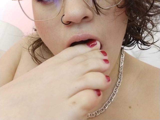 Foto de primer plano de Taylor, sin ropa, con cabello castaño rizado, un piercing en la nariz y gafas. Taylor, con las uñas pintadas de rojo, se muerde el dedo, llevando un collar de cadena gruesa y con la boca ligeramente abierta..