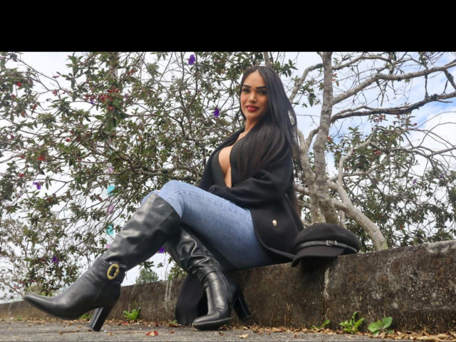 Vanezzattss está sentada en un borde de piedra, con las piernas separadas, botas negras con hebillas doradas, jeans azules, chaqueta negra, cabello oscuro. Sonrisa seductora, ramas de árboles en el fondo, entorno al aire libre, pose sensual..