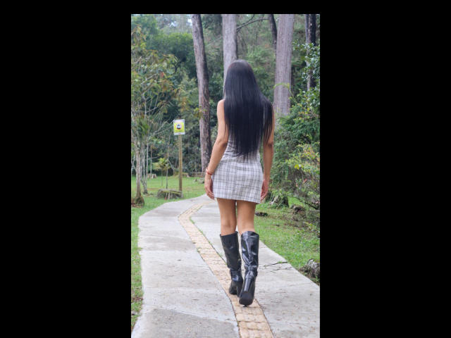 Vanezzattss, von hinten gesehen, geht einen Betonweg in einem Wald entlang. Sie hat langes, schwarzes Haar, trägt ein kurzes, kariertes Kleid und schwarze Stiefel, die bis zu den Oberschenkeln reichen. Ihr Hintern ist deutlich sichtbar..