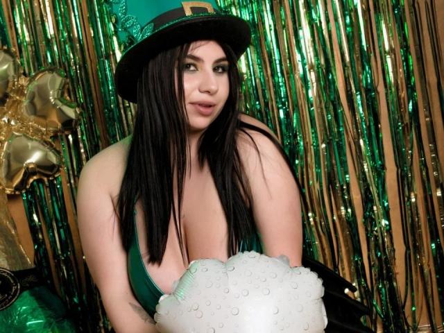 VenusxMusa, com cabelos longos e pretos e uma blusa verde, usa um chapéu preto com uma fivela dourada. Ela segura um adereço branco e fofo, com um fundo de cortina metálica verde e sexy, e um enfeite dourado em forma de estrela..