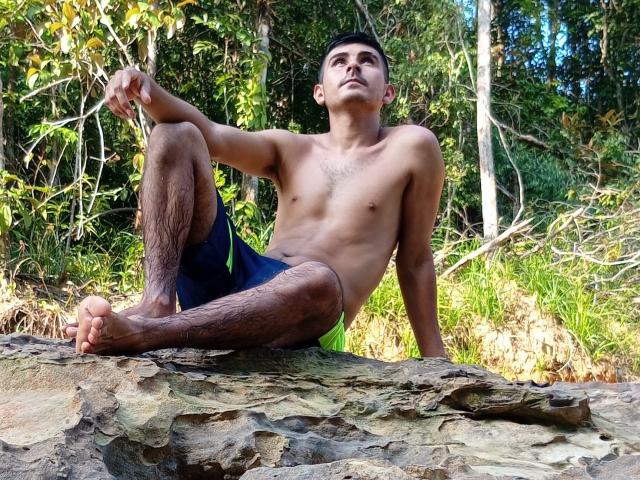 Charlly69, shirtless, sitting on rocky ground, legs spread, one arm behind, cock visible through dark shorts, forest background, sunlight, shadowed face, relaxed, sexy, natural light, outdoor, summer vibe.