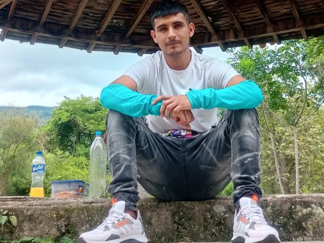 Charlly69, um homem atraente com cabelo preto curto, está sentado sob um telhado de madeira, com as pernas abertas, vestindo uma camiseta branca e mangas azuis, e calças pretas. calças jeans, tênis brancos, garrafa e pote ao fundo.