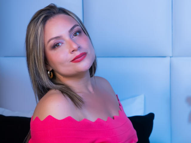 Foto van AshleyKonor met lichtbruin haar, rode lippenstift en een roze top met een open schouder, die recht in de camera kijkt. camera met een lichte kanteling, blauwe verlichting die haar huid accentueert. Seksuele spanning.