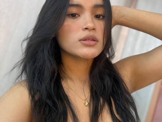 Gros plan de MiiaPalmer, une jeune femme aux longs cheveux noirs, aux yeux marrons et à la peau claire, posant avec son bras droit. Levé, regardant directement l'appareil photo. Elle porte un collier en or..