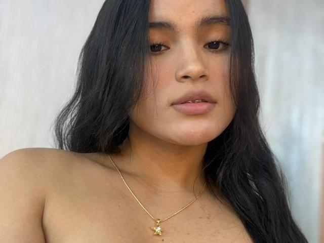 Primer plano de MiiaPalmer, una mujer latina con cabello negro largo, piel morena y labios carnosos, que lleva un collar dorado con colgante con forma de estrella pequeña. Está sin sujetador, mira directamente a la cámara, con los labios ligeramente entreabiertos..