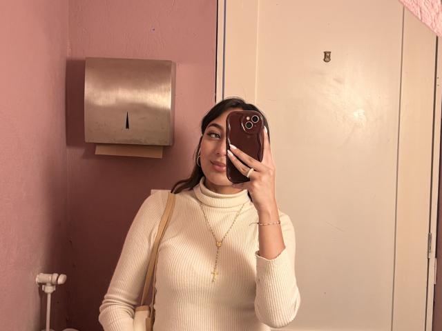 Fotografia di DianaFernandezz, una donna formosa con lunghi capelli scuri, che indossa un maglione a collo alto bianco a coste e tiene in mano uno smartphone, posizionato in un bagno con una parete rosa e un dispenser di carta da cucina in metallo. Si sta scattando un selfie allo specchio, con un'espressione sicura, con un sottile accenno di tensione sessuale..